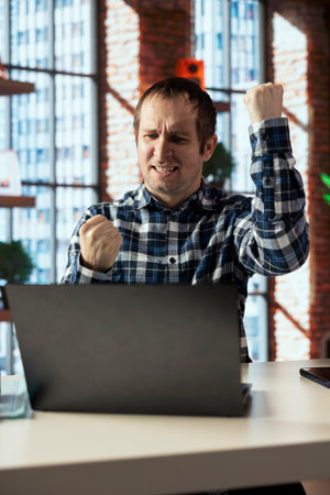 Joyous man at home office desk facing laptop screen, raising fists to celebrate an achievement. Overjoyed employee pumping fists in reaction to positive result at work, looking at laptop screenの写真素材
