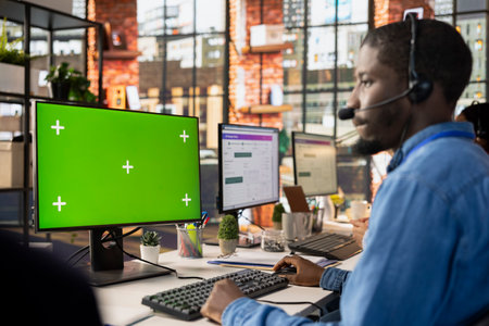 Call center worker in office solving customers issues using green screen PC. African american man in...