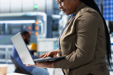 Black alternative energy consultant observes operations in a solar tech plant, ensuring factory practices meet green energy targets. Specialist uses laptop for manufacturing efficiency.の写真素材