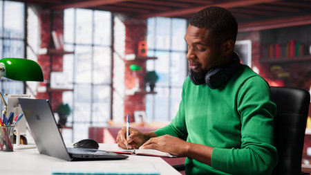 African american male teleworker writing personal notes at his desk in modern loft, remote job. Productive guy sitting at home office, using pen to take notes in his textbook. Camera A.の写真素材