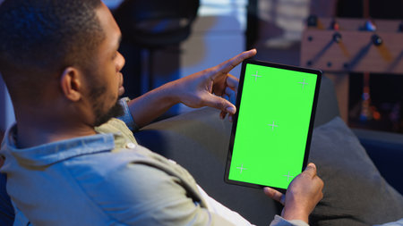 Relaxed male tech user holding device with isolated mockup screen in his living room, sitting on the couch and teleworking. African american happy guy browses the internet, outsourcing. Camera B.の写真素材