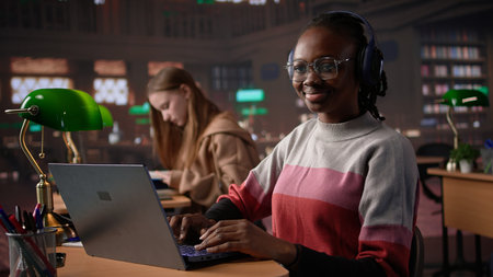 African american girl comparing course notes with data from library collection, using laptop to focus on a literature assignment. Young student working on her diploma project. Camera B.の写真素材