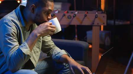 African American young adult working remotely on a laptop under loft neon lights, sitting on a couch in his cozy apartment. Relaxed person in a stylish home highlights modern productivity. Camera B.の写真素材