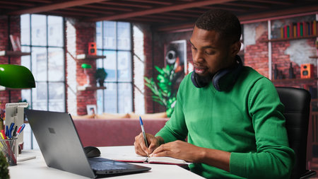 African american male teleworker writing personal notes at his desk in modern loft, remote job. Productive guy sitting at home office, using pen to take notes in his textbook. Camera B.の写真素材