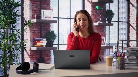 Professional woman in a modern loft takes a phone call at her desk. Multitasking with her laptop,...