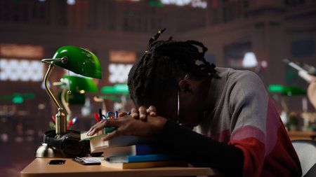 Female pupil struggling to stay focused and awake while researching, writing on her bachelor paper at the library in order to obtain her university diploma. Complete studies. Camera A.の写真素材
