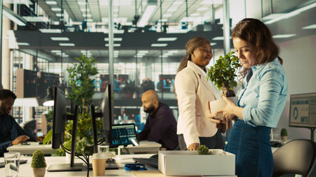 Employee getting consoled by colleagues after being fired, leaving office. Worker holding box filled with desk plants saying goodbye to coworkers after being laid off, camera Bの写真素材
