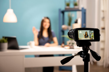 Behind the scenes of beauty vlogger filming tutorials, with a camera on a tripod focusing on her seated at a desk against a blue backdrop. Female content creator waving hello towards recording device.の写真素材