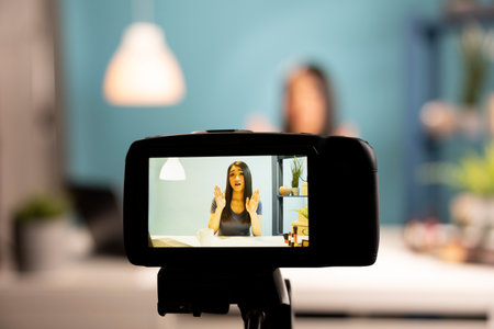 A digital camera capturing a beauty vlogger in action. Asian woman blogger is seen through the device screen, using expressive hand gestures while discussing makeup products in a studio setup.の写真素材