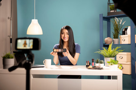 Female vlogger explains cosmetic products in a livestream video. Asian blogger holding eyeshadow palette, seated at a desk in studio with camera on tripod, creating engaging content for social media.の写真素材