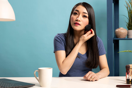 Professional makeup artist sits at her desk, demonstrating different brush techniques in a broadcasting session. Asian beauty content creator records a livestream focusing on cosmetic tools.の写真素材