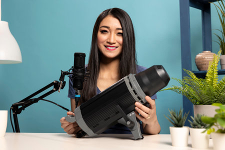 Asian woman holding a strobe light and speaking into a mic, presenting photography gear and accessories on her livestream. Female vlogger filming a product review video for her channel.の写真素材