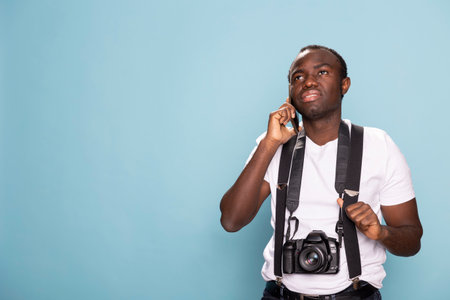 Young black man with a DSLR camera around his neck talking on his mobile device. Male photographer wearing suspenders and standing confidently in studio as he speaks with a client on his cellphone.の写真素材