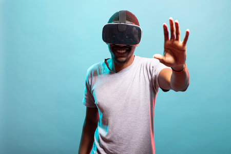 Smiling black man with his arm raised, wearing a VR headset, exploring a 3D simulation. African american male person enjoying a futuristic virtual reality experience with innovative technology.の写真素材