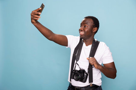 Black male photographer captures a selfie in a blue background studio, holding his cellphone confidently. He wears suspenders with a camera around his neck, exuding a stylish and modern look.の写真素材