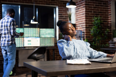 African american woman experiencing work fatigue massages her neck, taking a break from analyzing data on her laptop. The nighttime setting emphasizes the intensity of late shifts.の写真素材
