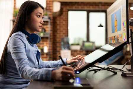 Asian graphic designer holding stylus pen, using digital art pad to modify images on computer screen. Skilled woman focused on retouching photos and refining design details for content production.の写真素材