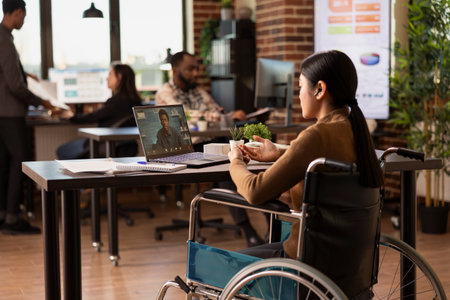 Female manager with a disability sitting in modern workspace, conducting a virtual meeting with a remote coworker. Asian woman in wheelchair with laptop, having a discussion with client on video call.の写真素材