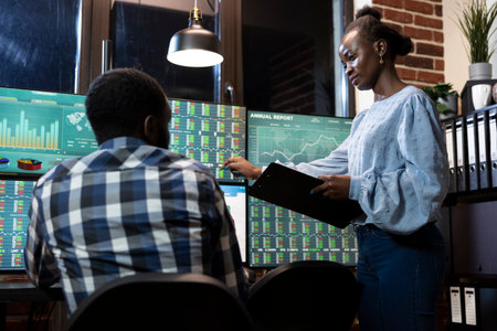 Two financial analysts collaborate at desk in office, reviewing stock market information on multiple monitors at night. Female manager with a clipboard, stands and explains forex trading to employee.の写真素材