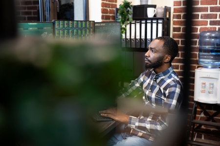 Focused financial expert seated at a desk, using digital technology during an online trading session. Black male entrepreneur analyzing stock market trends on multiple screens in a modern office.の写真素材