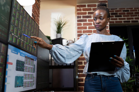 Stock analyst focuses on forex market at high tech workstation, tracking company hedge funds on multiscreen in brick wall office. Black woman with pen in hand, comparing data on screen and clipboard.の写真素材