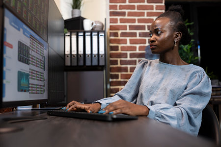 Focused black businesswoman staying up late at her desk, monitoring forex trading charts on several screens. Her brick wall office setting adds a modern touch to the work environment.の写真素材