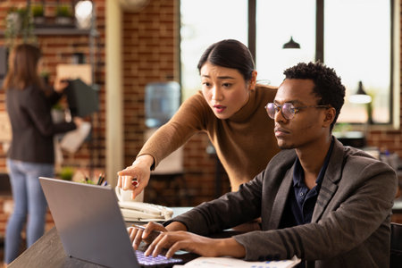 Female analyst points at laptop screen to explain market analysis to a african american coworker. Black man listens attentively as asian manager addresses financial errors in the presentation.の写真素材