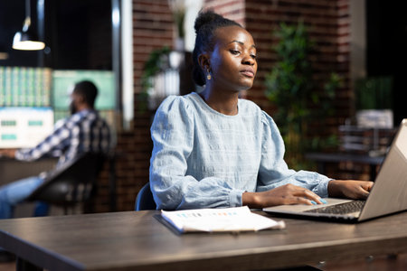 Young businesswoman with clipboard nearby, working late at night, analyzing financial data on her laptop. A blurred male coworker in the background focuses on multiple monitors.の写真素材