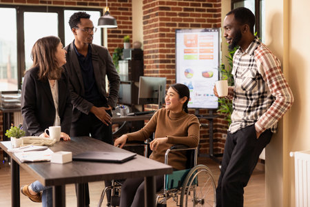 Multiethnic colleagues gather around a desk, taking a break and sharing stories in a relaxed office setting. Diverse group of employees stands, chatting with their asian coworker in a wheelchair.の写真素材