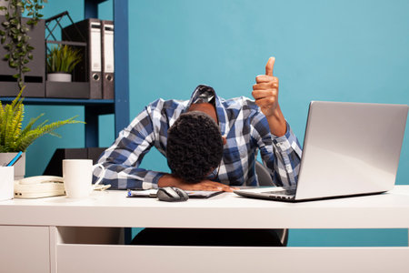 Young black businessman resting head on desk and giving thumbs up, exhausted but celebrating success. Sleepy office worker showing approval sign and napping on table after tiring workday.の写真素材