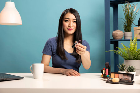 Asian beauty influencer seated at desk, filming a makeup tutorial. Female vlogger holds a lipstick, presenting it to camera and recording a video to engage her subscribers with expert cosmetic advice.の写真素材