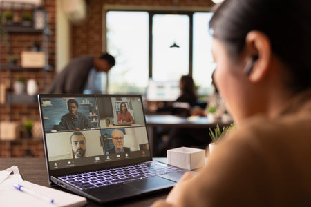 Selective focus on laptop displaying group of coworkers on a videoconference call with a businesswoman wearing earphones. Female manager sitting at desk, attending an online meeting with clients.の写真素材