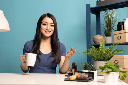 Young asian vlogger enjoying a cup of coffee while speaking to her viewers during a live session, filming in a studio with a blue background. The beauty influencer records content for her subscribers.の写真素材