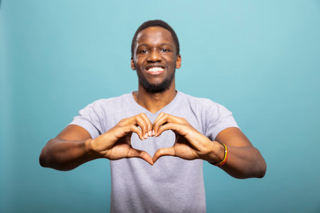 Charming young man making a heart with his hands, exuding warmth and joy against a blue background. African american male model posing with a love hand gesture, expressing affection in studio.の写真素材