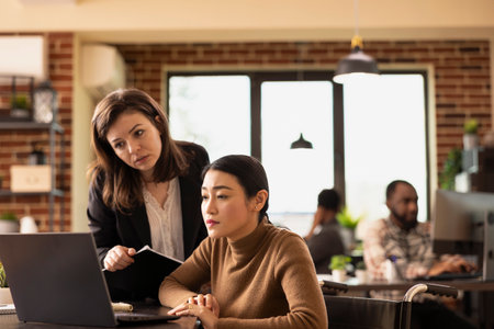 Caucasian woman with clipboard discusses marketing strategies with female colleague in wheelchair in brick wall office. Asian wheelchair user and her coworker looking closely at laptop screen.の写真素材