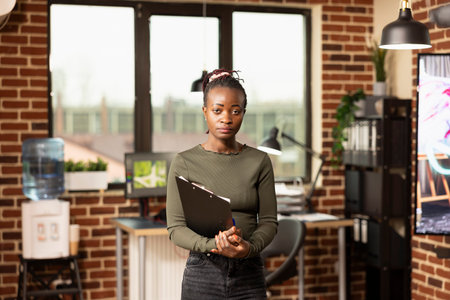 Young black woman confidently holding clipboard in brick wall office, ready to take on her role at creative agency. Professional african american agent standing in modern workspace, prepared for work.の写真素材
