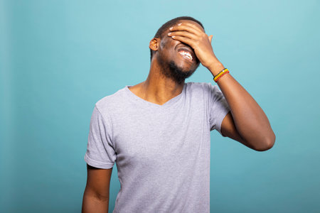 Cheerful male model covering part of his face, with a shy yet joyful expression against isolated backdrop. Portrait of black man laughing with hand on forehead, radiating warmth and positivity.の写真素材