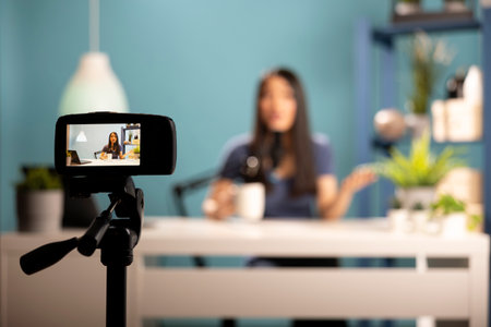 Asian woman blogger seated at desk in a professional studio setting, holding a cup of coffee and speaking into a microphone. The image emphasizes digital content creation and filming equipment.の写真素材