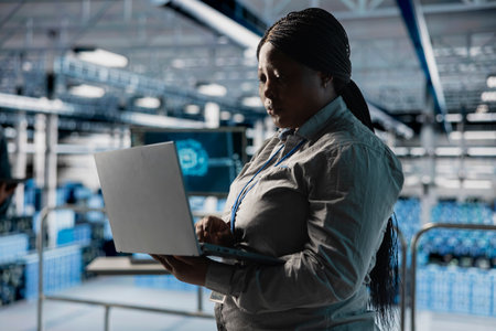 Data center female system administrator using laptop, configuring storage devices for minimal latency in AI workloads. African american woman using notebook in server farm to build neural networksの写真素材