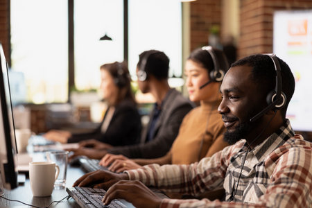 Closeup of african american call center agent reviewing information on his desktop pc while consulting with remote customers. Modern workspace with multiethnic telemarketers using their computers.の写真素材