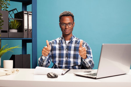 Portrait of african american man giving two thumbs up to camera. Young black guy seated at his desk with a laptop and clipboard, giving a supportive hand gesture of encouragement, an approval sign.の写真素材