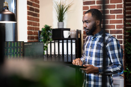 African american man with tablet, working night shift, examining financial reports on computer...