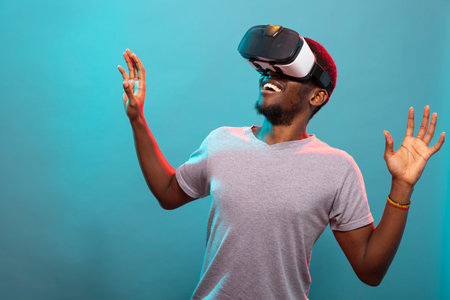 Cheerful young man using VR goggles, standing with his arms raised against a blue background. African american guy with a surprised expression, experiencing an interactive 3D virtual simulation.の写真素材