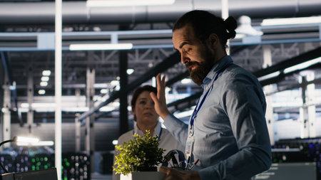 Furious data center engineer packs desk belongings after being replaced by AI automation handling operations. Frustrated man suffering from workforce displacement by artificial intelligence, camera Aの写真素材