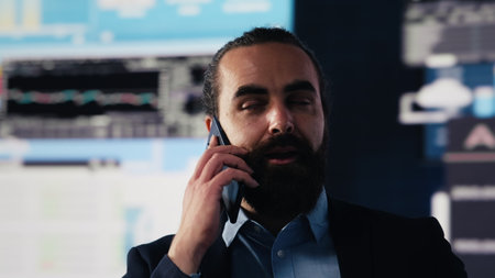 Businessman answering a smartphone call to evaluate digital analytics and corporate insight. Surrounded by finance data and trend reports, he strategizes leadership goals and projections. Camera A.の写真素材