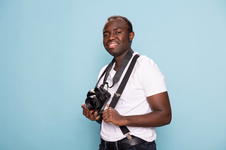 African american photographer posing with a relaxed yet confident expression, gripping his suspenders and holding his camera. The solid blue background adds a modern and clean look.の写真素材