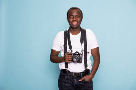 Black photographer posing with a camera around his neck, exuding professionalism. He stands against a blue background, highlighting his casual fashion and creative essence.の写真素材
