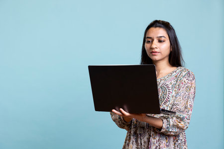 Indian young adult composing email or typing on her laptop in studio, solving her professional tasks on pc. Casual focused person browses web online standing against blue background.の写真素材