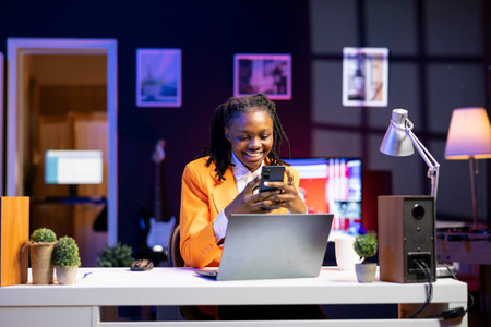 Happy woman at home having fun looking at social media reels done by online content creators. Smiling african american teenager relaxing by using mobile social network app at laptop deskの写真素材