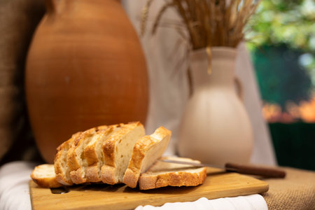 A sliced loaf of bread is on a wooden cutting board. A large clay pot and a white vase are in the...